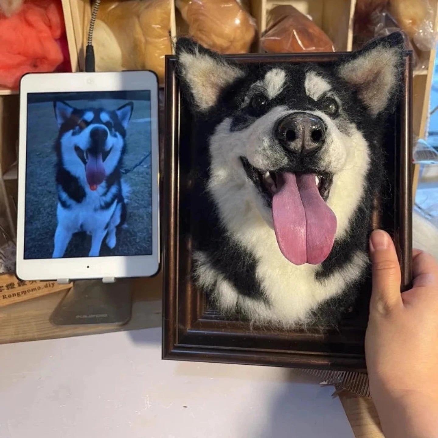 Realistic handmade wool felt portrait of a dog displayed in a wooden frame beside its reference photo